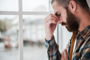 Feeling hopeless. Depressed young bearded man keeping eyes closed and touching his face while standing near the window